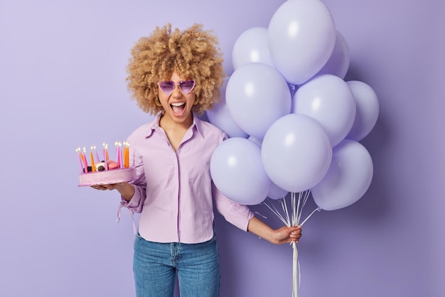 Festive occasion and celebration concept Emotional woman with curly hair holds sweet cake and bunch of helium balloons exclaims loudly wears shirt jeans and sunglasses isolated over purple wall