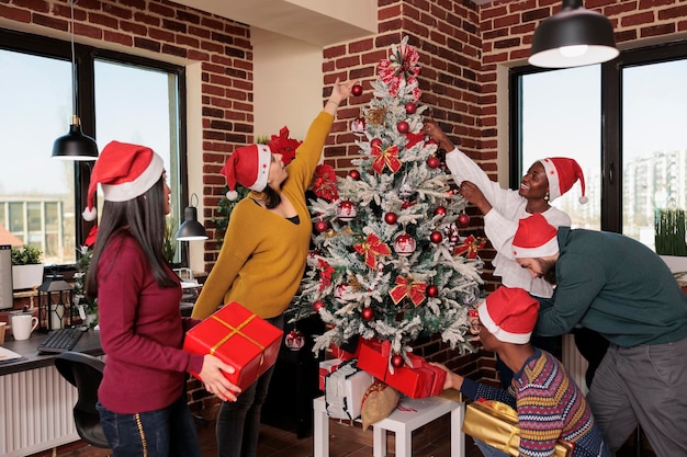 Foto gente festiva che fa regali sotto l'albero di natale nell'ufficio dell'azienda, celebrando le vacanze invernali al lavoro. scambio di regali per la tradizione natalizia sul posto di lavoro con decorazioni stagionali.