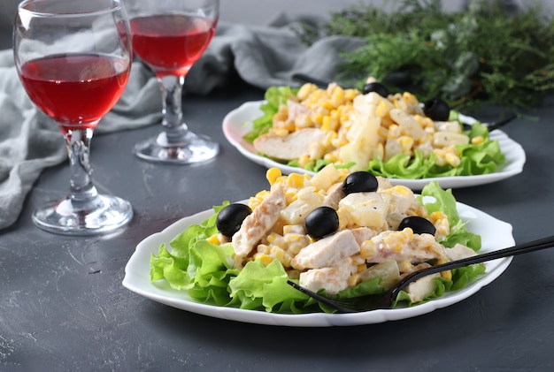 Festive salad with pineapple, baked chicken, corn and black olives on white plates and two glass of red wine on grey table. Horizontal format