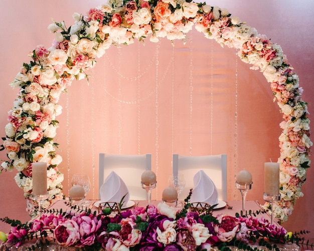 festive table arch stands decorated with composition of violet purple pink flowers and greenery candles in the banquet hall table newlyweds in the banquet area on wedding party