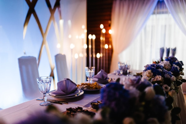 Festive table decorated with composition of flowers and
greenery candles in the wedding banquet hall table newlyweds
covered with a tablecloth and served with dishes and cutlery close
up