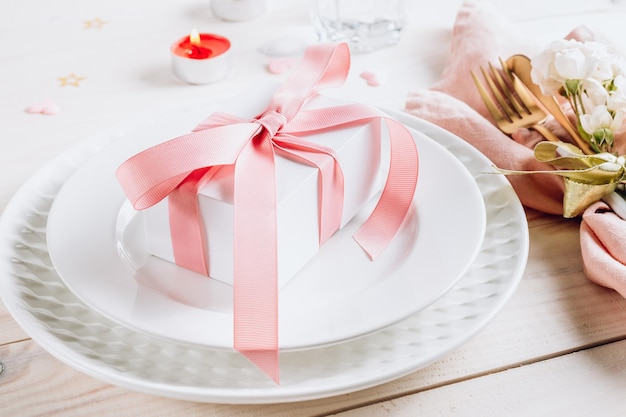 お祝いのテーブルセッティング、白い木製の背景にピンクのナプキンとギフトボックスが付いた皿とカトラリー、美しい配置、選択と集中