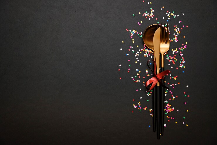 Festive table setting for Valentine's Day with fork, knife and hearts on a black table with confetti. Space for text.