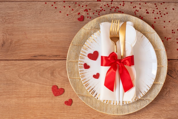 Festive table setting for Valentine's Day with fork, knife, red bow and hearts on a wooden table. Space for text. Top view