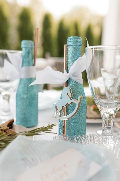 Праздничная сервировка стола с украшенной блестящей синей бутылкой напитка на белой скатерти крупным планом деко