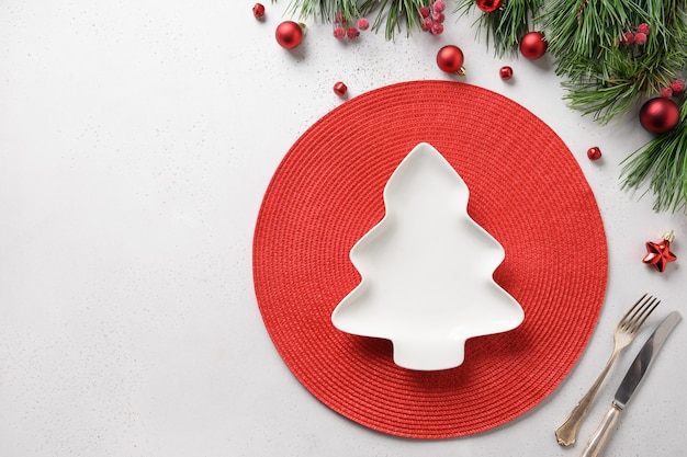 Сервировка праздничного стола с тарелкой в форме рождественской елки на белом фоне с красным украшением. Вид сверху. Место для текста.