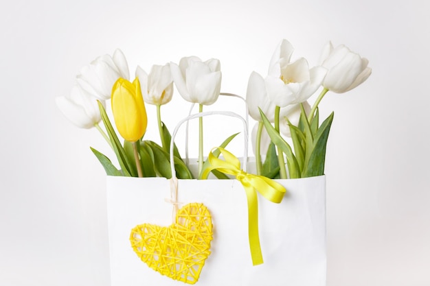 Festive white yellow tulips composition handmade heart ribbon on white background Bouquet of spring flowers in white paper bag Copy space Birthday Mothers Valentines Womens Day concept