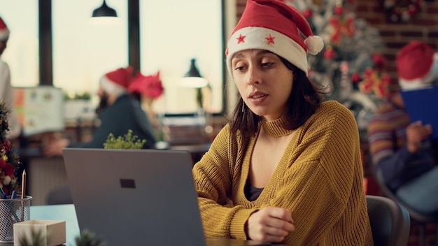 Festive woman talking on online videocall conference, attending teleconference meeting with webcam during christmas holiday festivity. Chatting on videoconference in xmas decorated office.