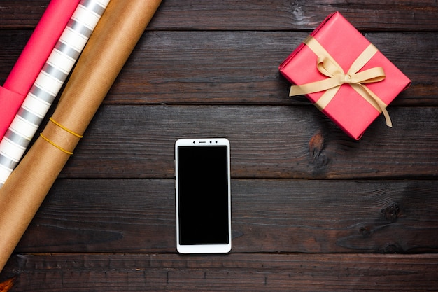 Праздничная упаковочная бумага, розовый подарок и белый телефон на темном фоне. Вид сверху.