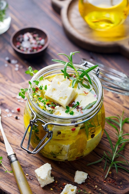 Feta cheese marinated in olive oil with fresh herbs in glass jar Wooden background
