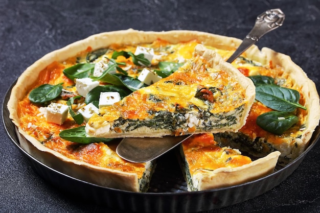 Feta spinach cheddar cheese quiche in a baking dish on a wooden board on a grey concrete table with a slice on a spatula, french cuisine