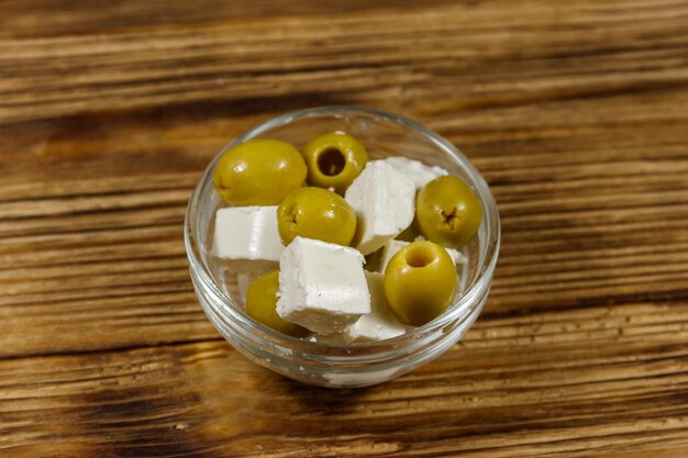 Fetakaasblokjes met groene olijven in glazen kom op een houten tafel