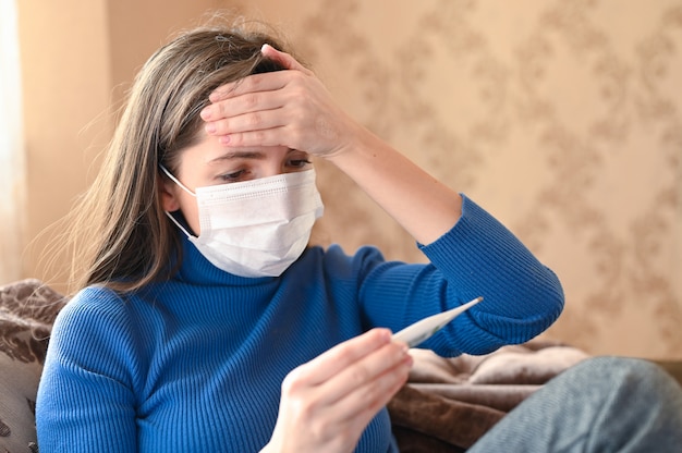 Fever and coronavirus symptoms, woman in medical mask measures body temperature. Close-up