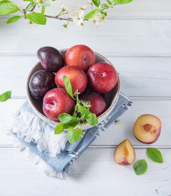 A few juicy red plums in bowl  with branch blossom flowers on a white  wood background. Top view and copy  space  image