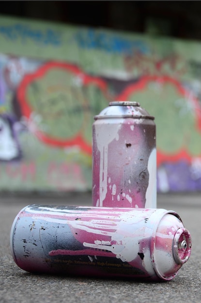 Alcune bombolette spray usate con vernice rosa e bianca giacciono sull'asfalto sullo sfondo di un muro dipinto in coloratissimi disegni di graffiti