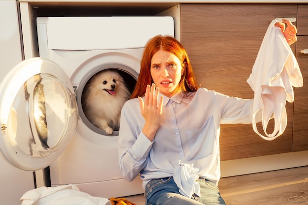 なんて悪臭だ！ショックを受けた興奮した赤毛の女の子が汚れた臭いの服の山を洗う、彼女の愛らしいふわふわのスピッツhelpinhは自宅の洗濯機の中にあります