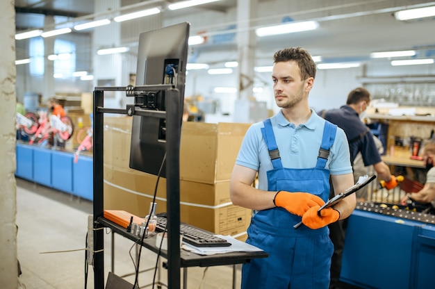 Fietsfabriek, werknemer met laptop vormt op de assemblagelijn van de fiets. Mannelijke monteur in uniform installeert fietsonderdelen in de werkplaats