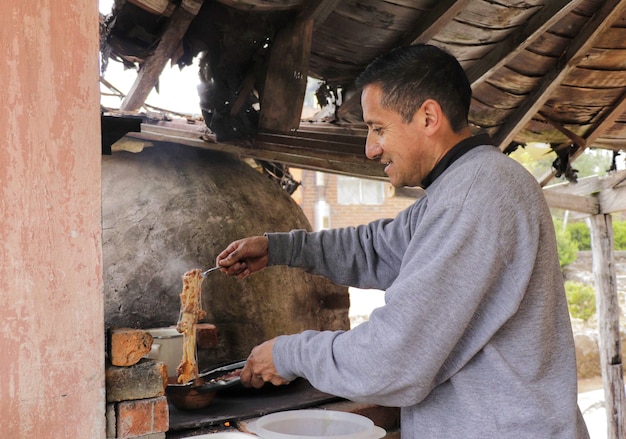 Fifty year old adult man cooking cheese in a small clay pot on\
a wood stove in an outdoor kitchen under a wooden kiosk wearing a\
gray sweatshirt