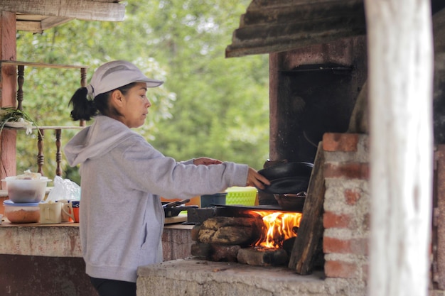 Fifty year old adult woman cooking using a frying pan and a\
small clay pot in an outdoor wood-burning kitchen with a foliage\
background in a gray sweatshirt and cap