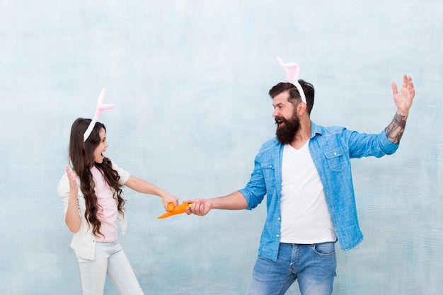 Fight for prize easter carrot fight happy family wear bunny ears fun and happiness father and daughter hold carrot spring holiday party small kid girl and dad in rabbit costume healthy food