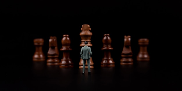 Figures businessman standing in front of wooden chess on black isolated background.