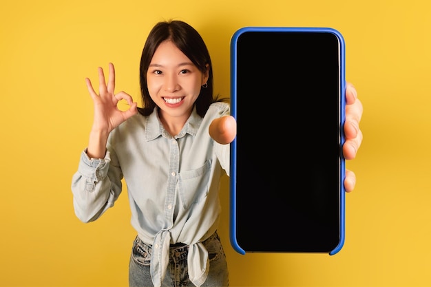 Fijne aantrekkelijke aziatische vrouw met een gloednieuwe smartphone met een leeg scherm waarop een goed gebaar te zien is