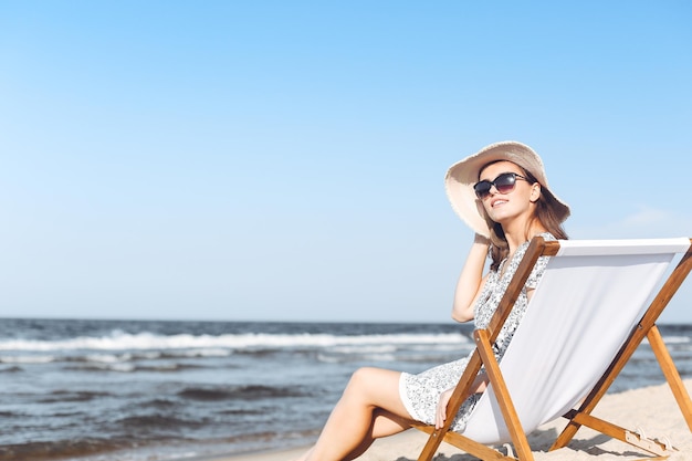 Fijne brunette vrouw met zonnebril en hoed ontspannend op een houten ligstoel op het oceaanstrand.