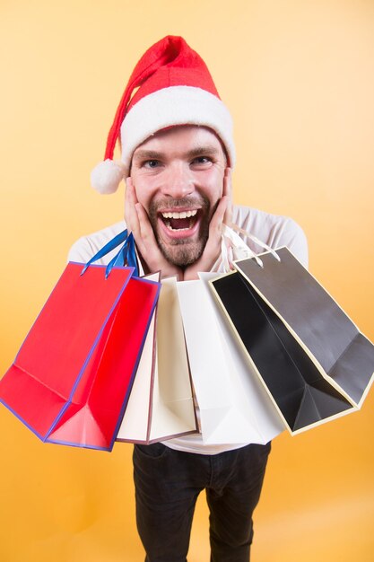 Fijne feestdagen viering. Man in kerstmuts met zakken op oranje achtergrond. Kerstmis, nieuwjaarsverrassing, cadeautjes. Macho shopper met verbaasd gezicht houdt paperbags vast. Winterverkoop, winkelconcept.