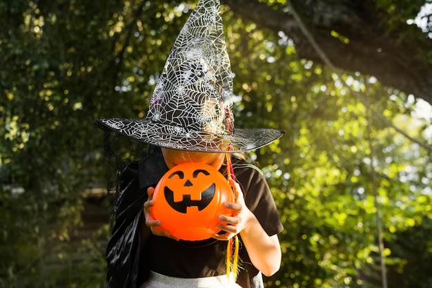 Fijne Halloween-dag Klein meisje gekleed in een heksenkostuum houdt een emmer snoep vast en kijkt naar snoep