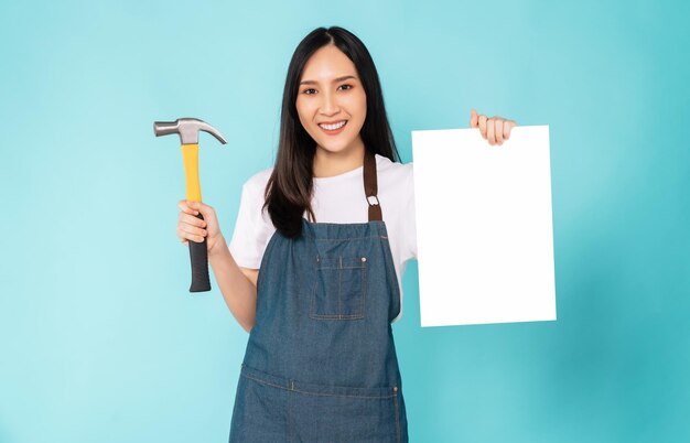 Fijne jonge Aziatische vrouw draagt een schort en houdt een timmermanshamer vast met een blanco papier op een blauwe achtergrond