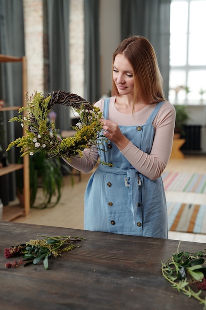 Fijne jonge blonde vrouw in vrijetijdskleding die bij een houten tafel in een huiselijke omgeving staat en de paaskrans voorbereidt op vakantie