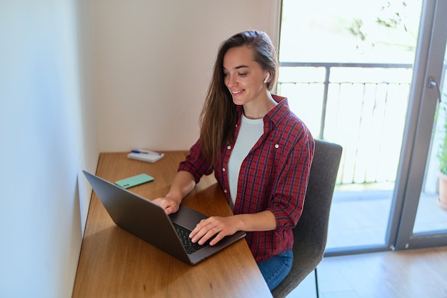 Fijne jonge, casual, slimme, moderne lachende meisjesfreelancer met draadloze hoofdtelefoon die een laptop gebruikt tijdens thuiswerken op afstand