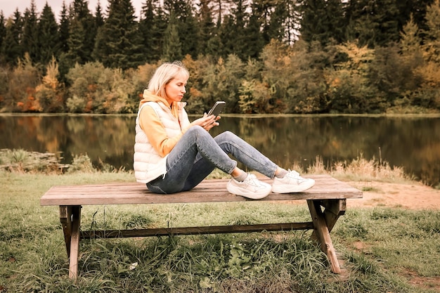 Fijne jonge mooie vrouw die buiten in het herfstpark loopt met behulp van een mobiele telefoon die een schilderachtig uitzicht op de rivier heeft