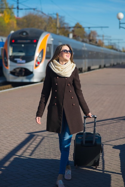 Fijne jonge vrouw met een bril gekleed in een jas met grote reistas op het treinstation op de treinachtergrond