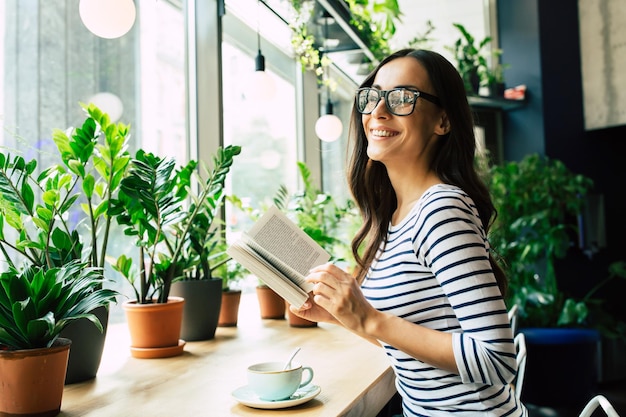 Fijne mooie jonge vrouw met een bril leest een boek en drinkt koffie in een modern café