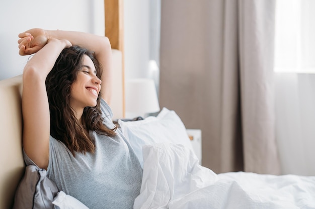 Fijne ochtend Thuisontspanning Positieve levensstijl Tevreden vrolijke brunette vrouw die alleen geniet van rust in een gezellig bed glimlachend in lichte slaapkamer