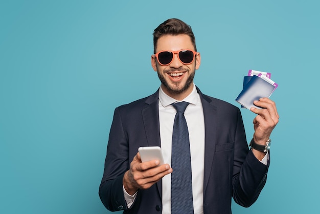 Fijne zakenman in een zonnebril die een smartphone gebruikt en naar de camera glimlacht terwijl hij paspoorten en