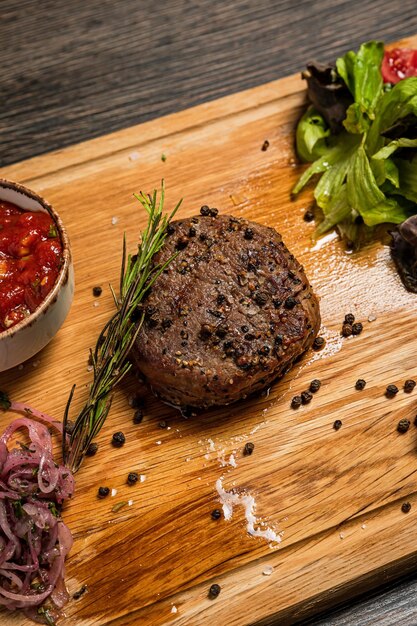 Photo filet mignon steak vertival photo closeup served on wooden board with spicy tomato sauce fresh vegetable salad pickled onions and rosemary