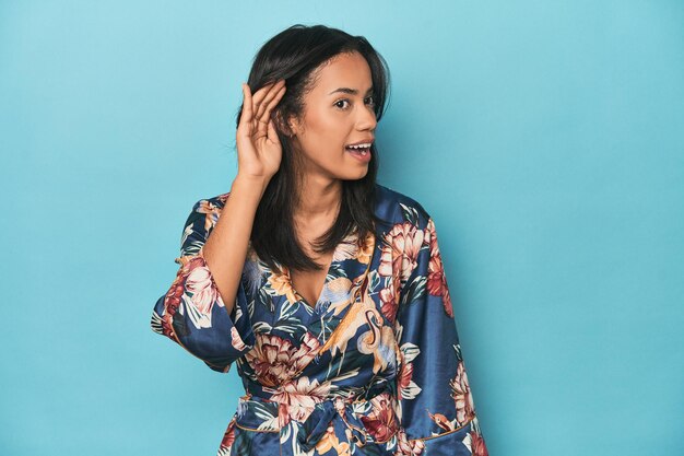 Filipina in floral kimono on blue studio trying to listening a gossip