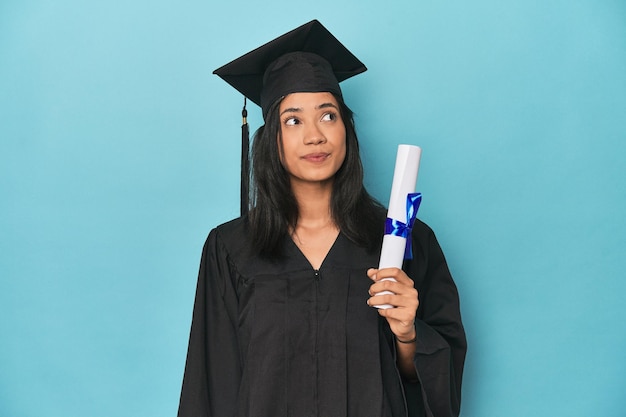 Photo filipina graduate with diploma on blue studio dreaming of achieving goals and purposes