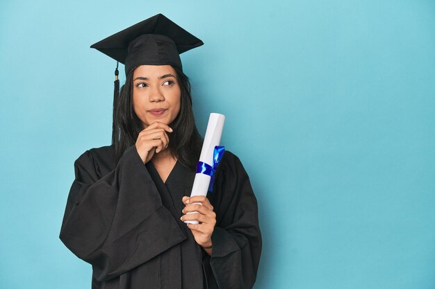 Photo filipina graduate with diploma on blue studio looking sideways with doubtful and skeptical