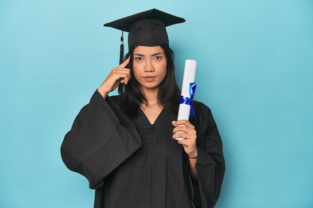 Photo filipina graduate with diploma on blue studio pointing temple with finger thinking focused on a task