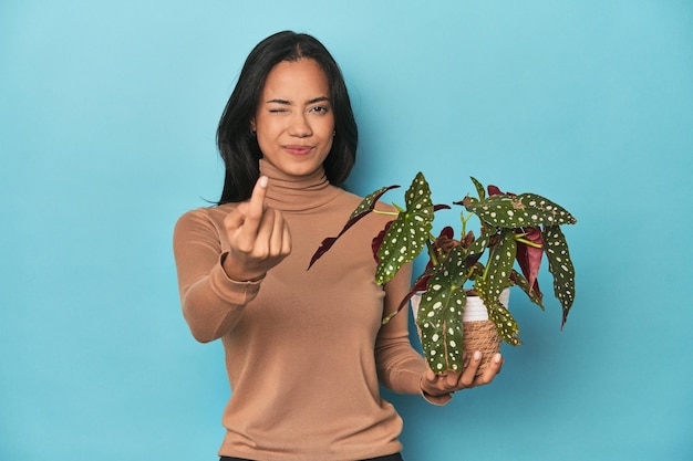 Filipina holding a plant on blue studio pointing with finger at you as if inviting come closer