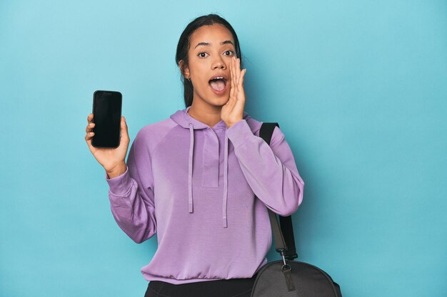 Filipina ready for gym with phone on blue shouting and holding palm near opened mouth