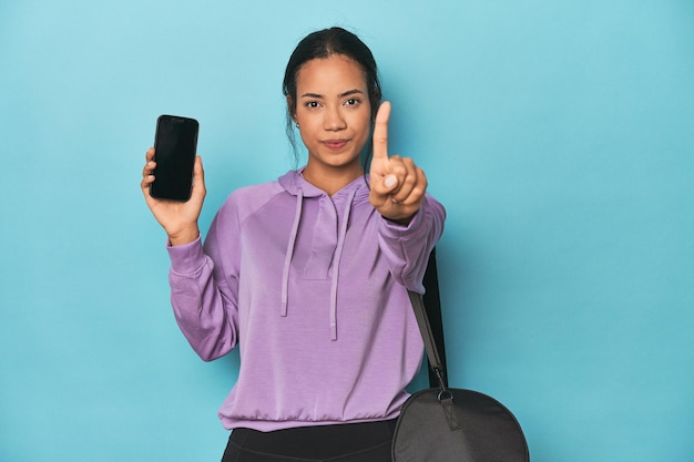 Filipina ready for gym with phone on blue showing number one with finger