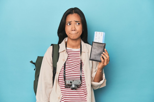 Photo filipina with camera tickets backpack on blue confused feels doubtful and unsure