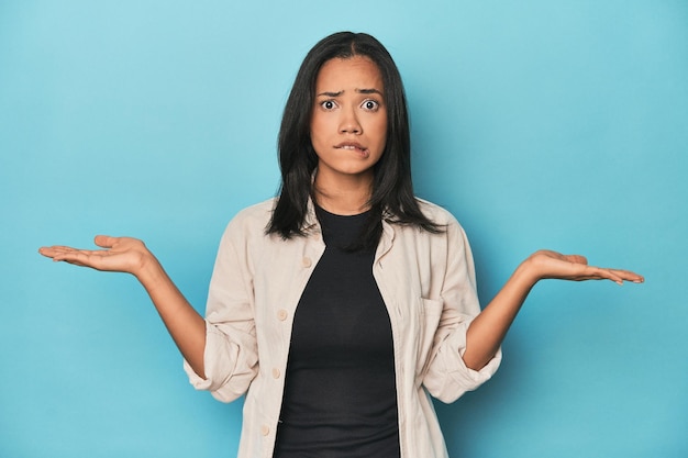 Filipina young woman on blue studio confused and doubtful shrugging shoulders to hold a copy space