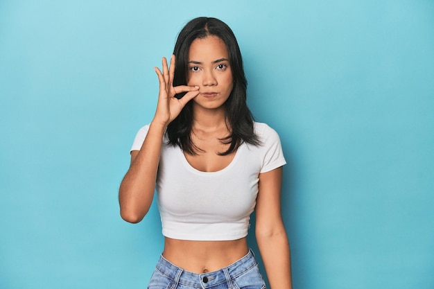 Filipina young woman on blue studio with fingers on lips keeping a secret