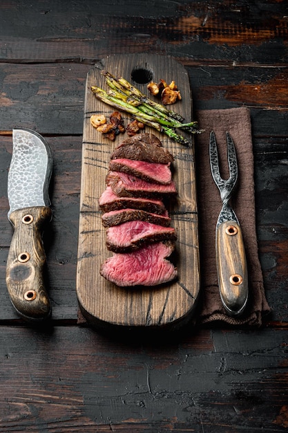 Filetto filetto mignon bistecche di manzo grigliate e affettate con cipolla e asparagi su tavola di legno con coltello da carne e forchetta su sfondo vecchio tavolo in legno scuro