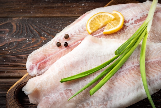 Filetto di pesce pangasio crudo con limone e cipolla verde su tagliere su sfondo scuro.